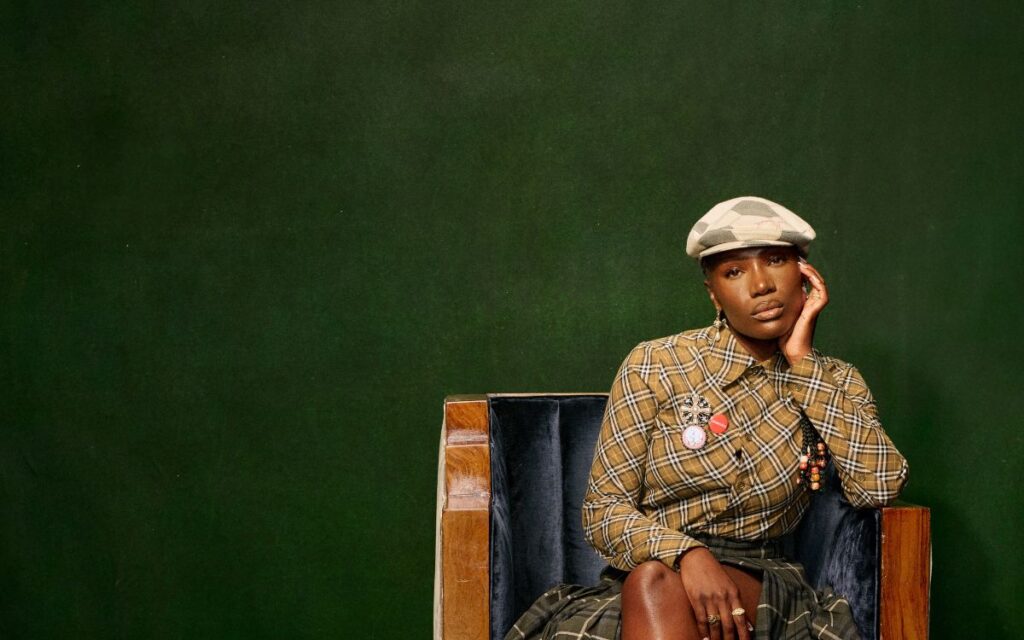 American rapper, singer, songwriter, Doechii. She's wearing a mustard plaid jacket and seated in a wood and velvet chair seated against a dark green wall