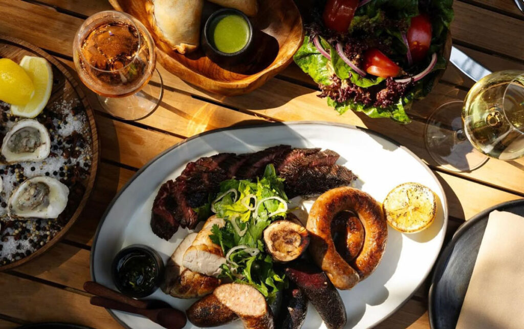 An overhead perspective of a meal featuring steak, sausage, oysters, salad, and cocktails at Argentinian restaurant, Porzana, located in the Minneapolis North Loop neighborhood.