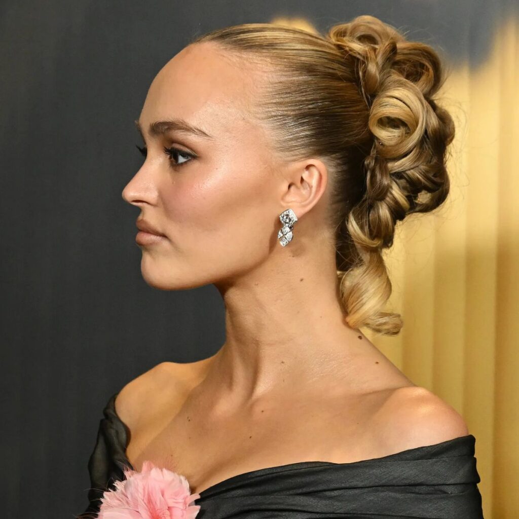 Lily-Rose Depp wearing an off the shoulder gown for a special event. Her long hair is pulled back with dramatic sculptured curls.